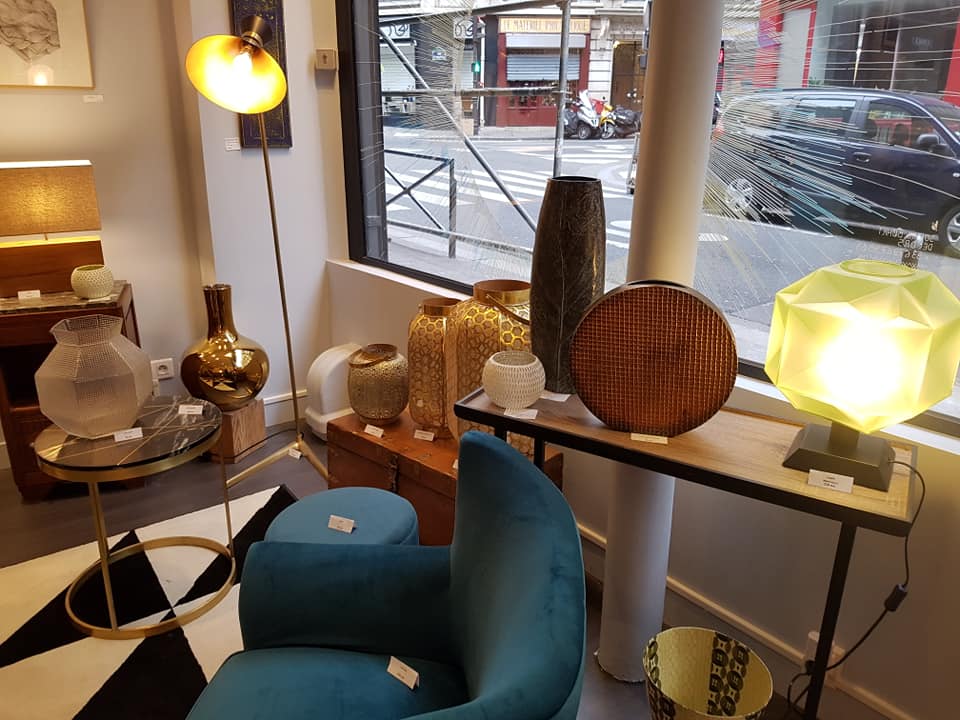 Intérieur de boutique d'ameublement moderne avec lampadaire doré, fauteuil bleu sarcelle, et une console près de la fenêtre montrant la rue urbaine.